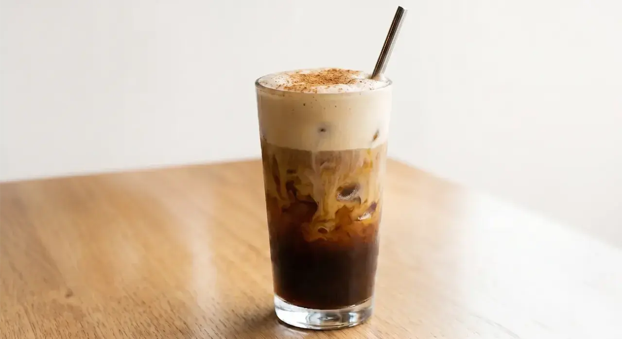 Ein erfrischender, geschüttelter Espresso mit cremiger Haube und Zimt bestreut, serviert in einem Glas mit Eiswürfeln und einem Metallstrohhalm.