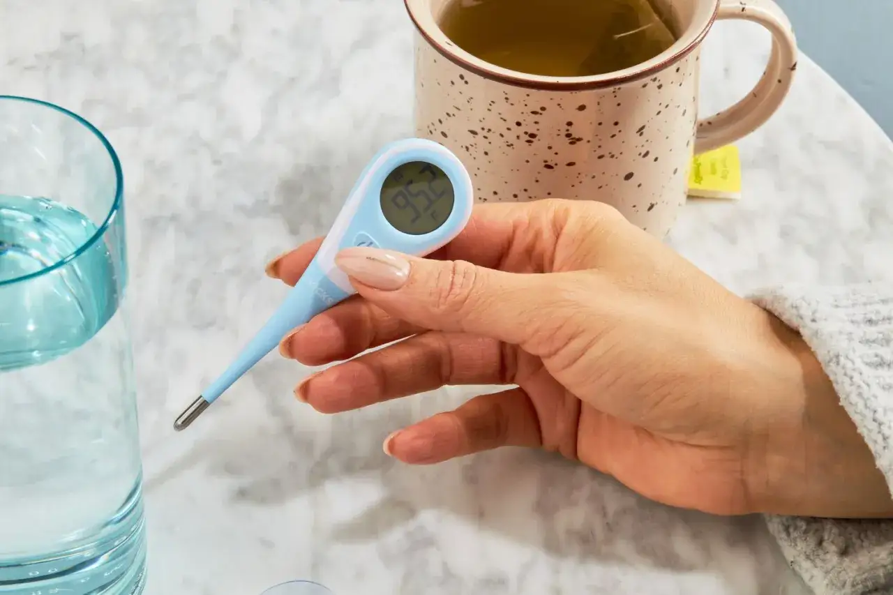 Digitale Fieberthermometer-Messung, während eine Tasse Tee daneben steht. Kaffee bei Fieber ist vielleicht nicht ideal, aber ein Tee kann helfen.