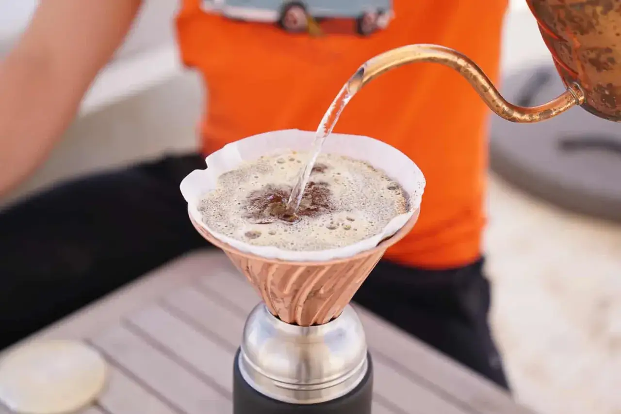 Hei&szlig;es Wasser flie&szlig;t aus einer Kupferkanne in einen Papierfilter mit Kaffeepulver. So entsteht k&ouml;stlicher Filterkaffee.