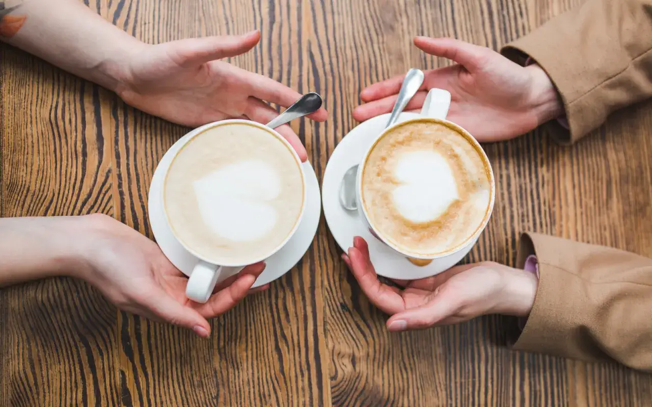 Zwei H&auml;nde reichen sich zwei Tassen mit Milchschaum-Herz. Ein Milchkaffee oder Cappuccino f&uuml;r jeden.