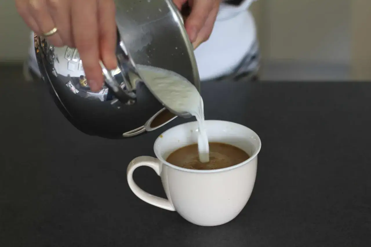 Milch wird aus einem Metallk&auml;nnchen in eine Tasse Kaffee gegossen. Perfekte Milchkaffee Zubereitung.
