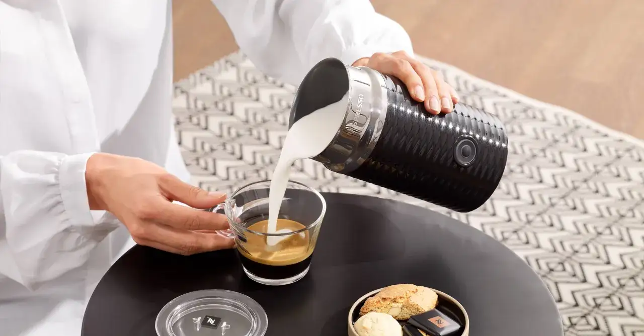 Perfekte Cappuccino Zubereitung: Milch wird aus einem Milchaufschäumer in einen Espresso gegossen. Daneben liegen Kekse und Schokolade.