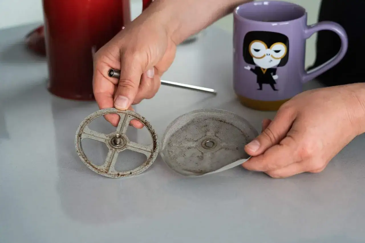 H&auml;nde zerlegen eine schmutzige French Press, um sie zu reinigen. Kaffeereste sind auf dem Metallfilter und dem Rahmen sichtbar.
