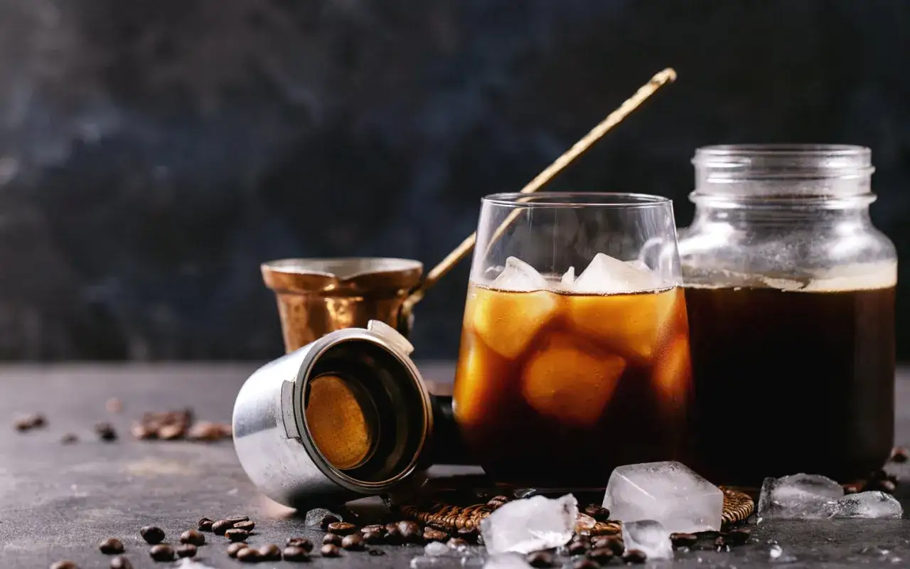 Ein Glas mit Eisw&uuml;rfeln und Cold Brew Kaffee, daneben ein Glasbeh&auml;lter mit Kaffee. Die Frage 