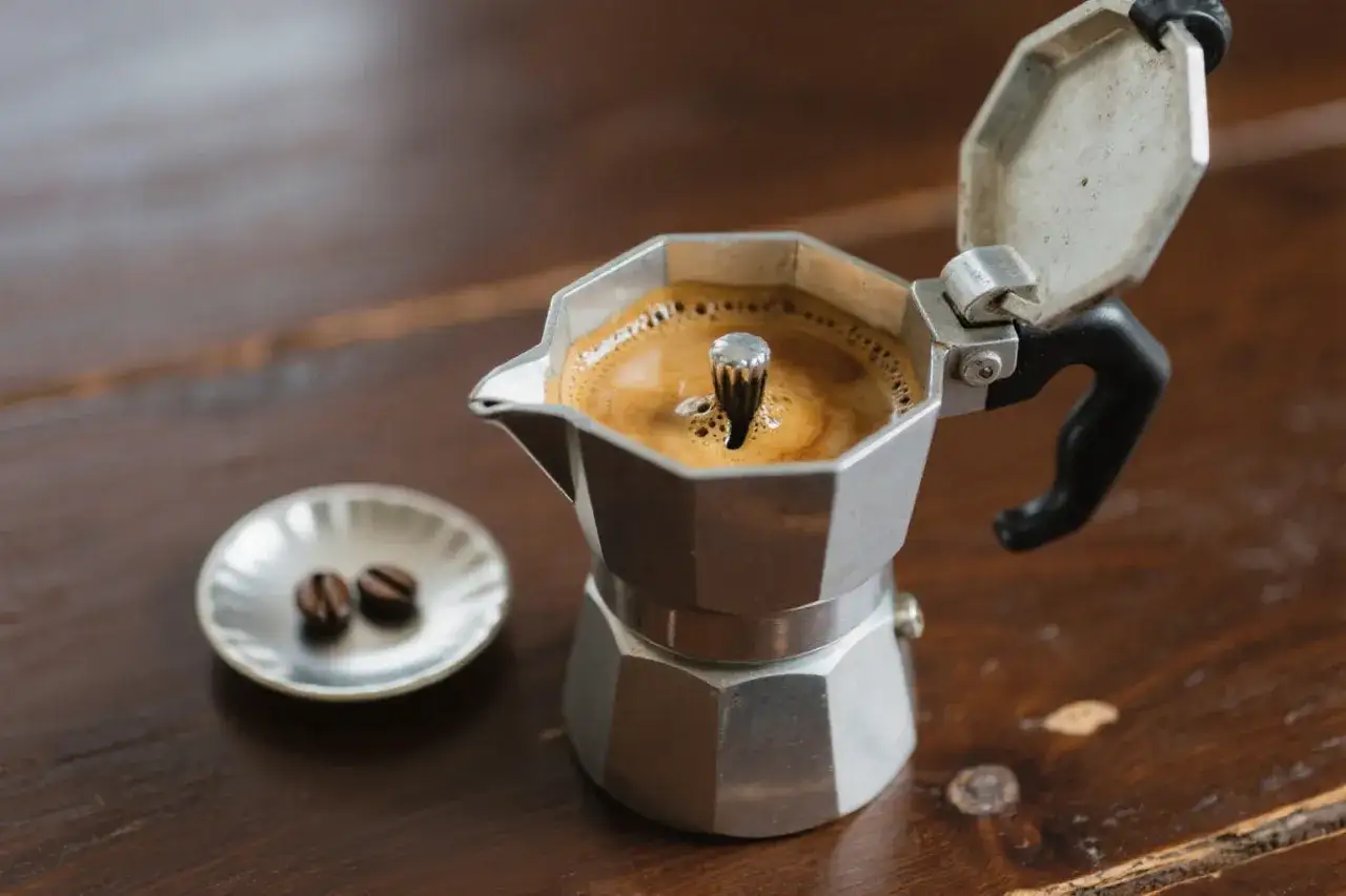 Frisch gebr&uuml;hter Kaffee auf dem Herd in einer Mokkakanne, daneben zwei Kaffeebohnen auf einem kleinen Teller.