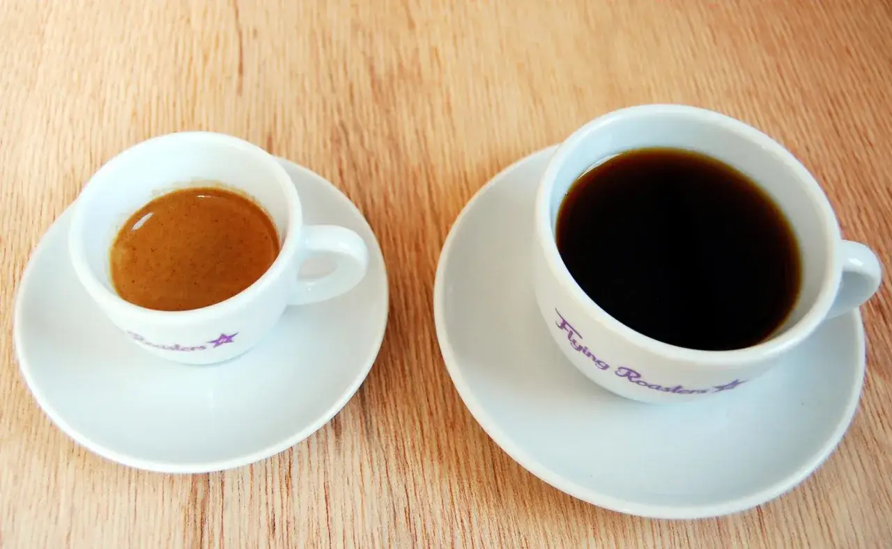 Zwei Tassen auf Holzhintergrund: ein kleiner Espresso mit Crema und ein größerer schwarzer Kaffee. Der Unterschied zwischen Espresso und Kaffee wird hier deutlich.