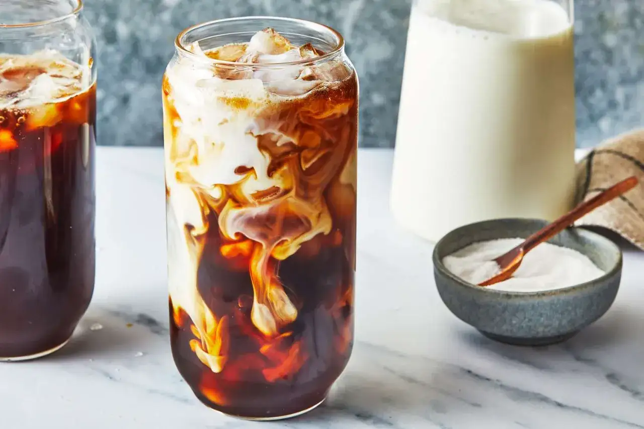Ein erfrischender Eiskaffee ohne Eis, mit cremiger Milch, die sich in dunklem Kaffee wirbelt. Daneben eine Zuckerschale.
