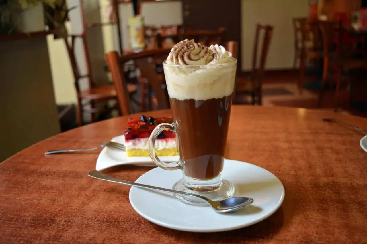 Ein Glas mit braunem Kaffee, gekr&ouml;nt von Sahne und Kakaopulver, steht auf einem Tisch neben einem St&uuml;ck Kuchen.
