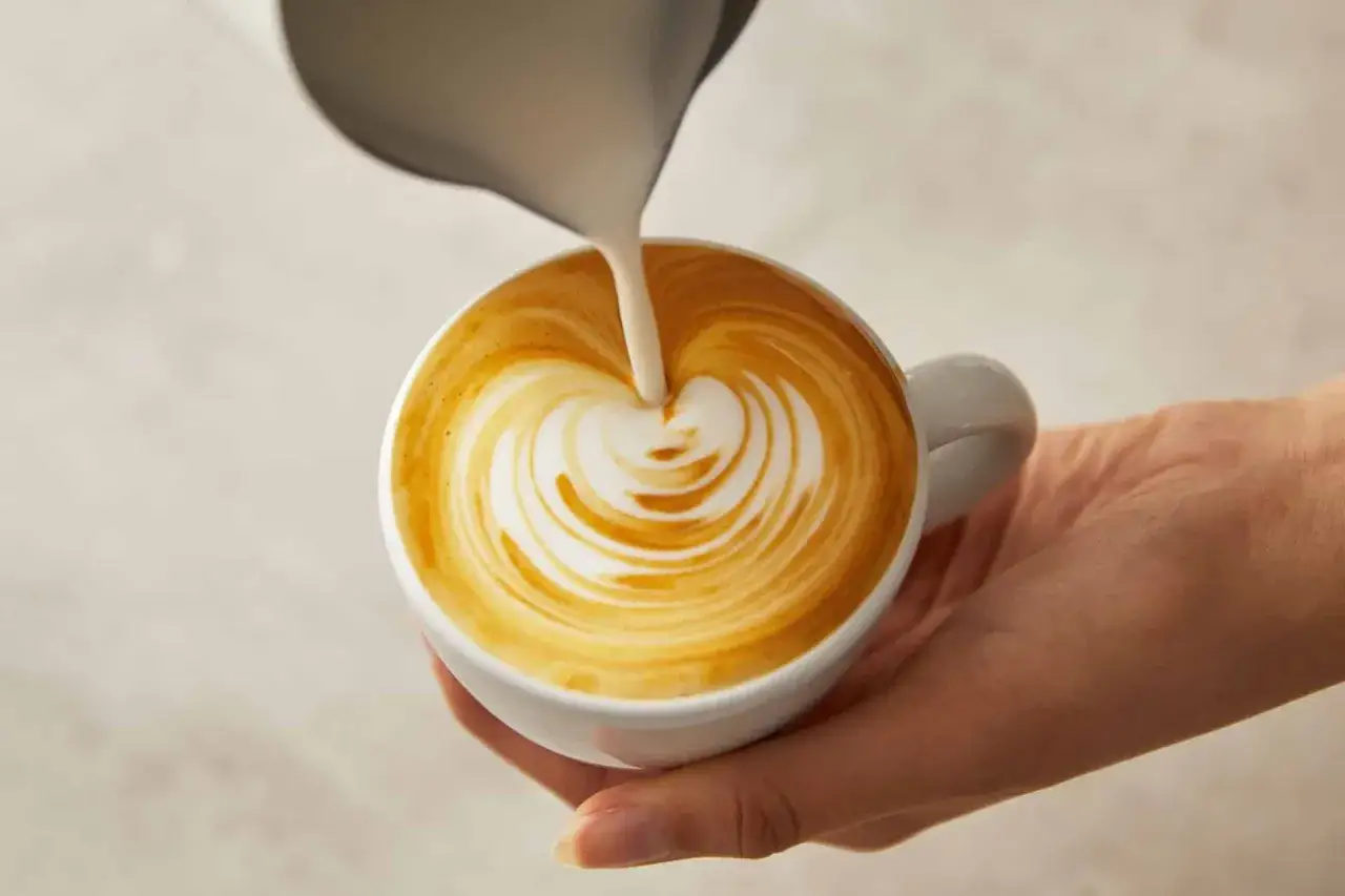 Eine Hand hält eine Tasse Kaffee, in die gerade Mandelmilch gegossen wird. Ein Herzmuster entsteht auf dem Schaum.