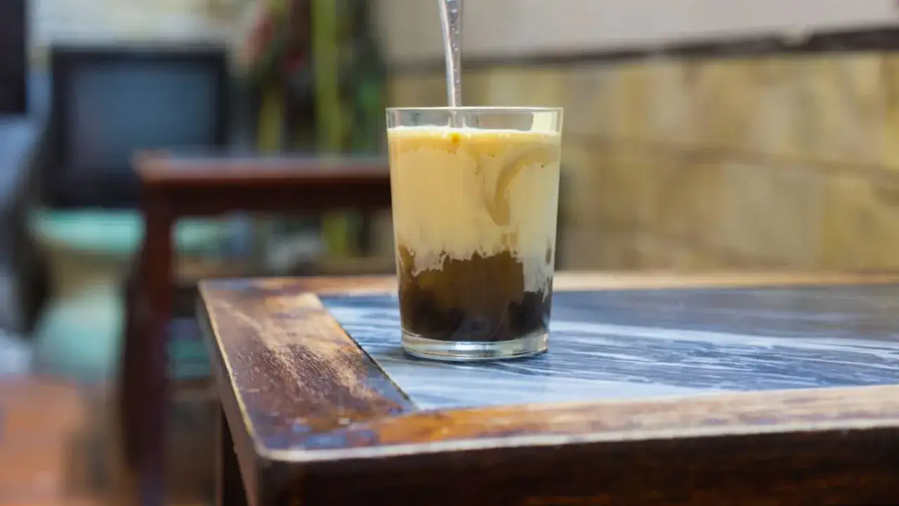 Ein Glas mit geschichtetem Kaffee mit Ei steht auf einem Holztisch. Die cremige obere Schicht vermischt sich mit dem dunklen Kaffee darunter.