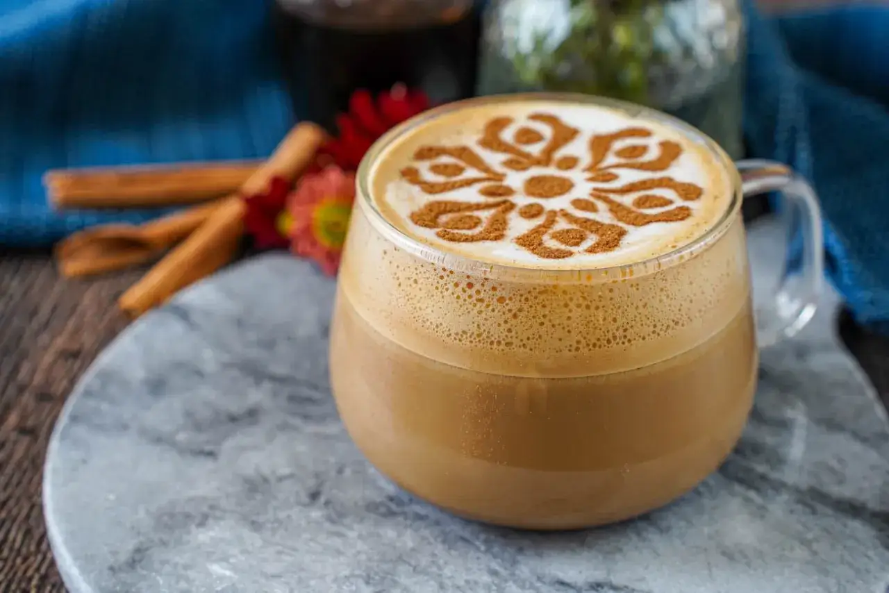 Ein köstlicher Zimt-Latte mit einem kunstvollen Zimtmuster auf dem Milchschaum, daneben Zimtstangen und Blumen.