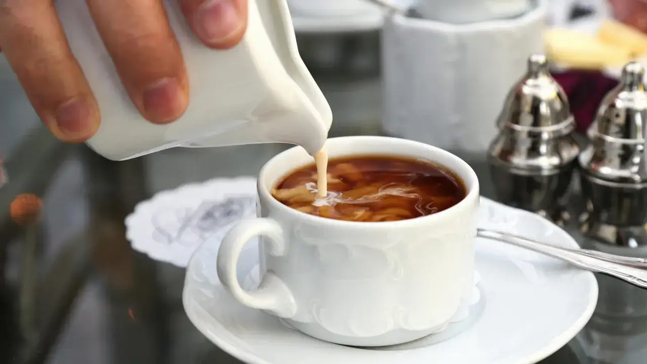 Cremiger Guss in eine Tasse Kaffee. Ein kleiner Krug gibt Milch in den hei&szlig;en Kaffee, was den Unterschied zwischen einem Americano und einem Kaffee mit Milch zeigt.