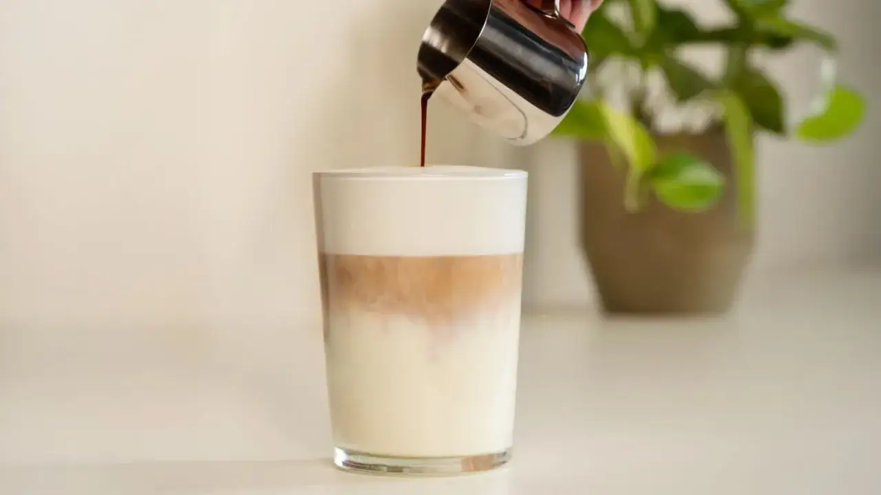 Ein Latte Macchiato wird in ein Glas gegossen. Die Schichten aus Milch, Kaffee und Schaum sind sichtbar. Weniger Latte Macchiato Kalorien mit fettarmer Milch.