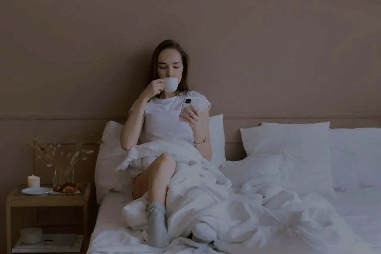 Eine junge Frau sitzt im Bett, genießt ihren Kaffee und scrollt durch ihr Handy. Ein gemütlicher Abend, um abzuschalten.