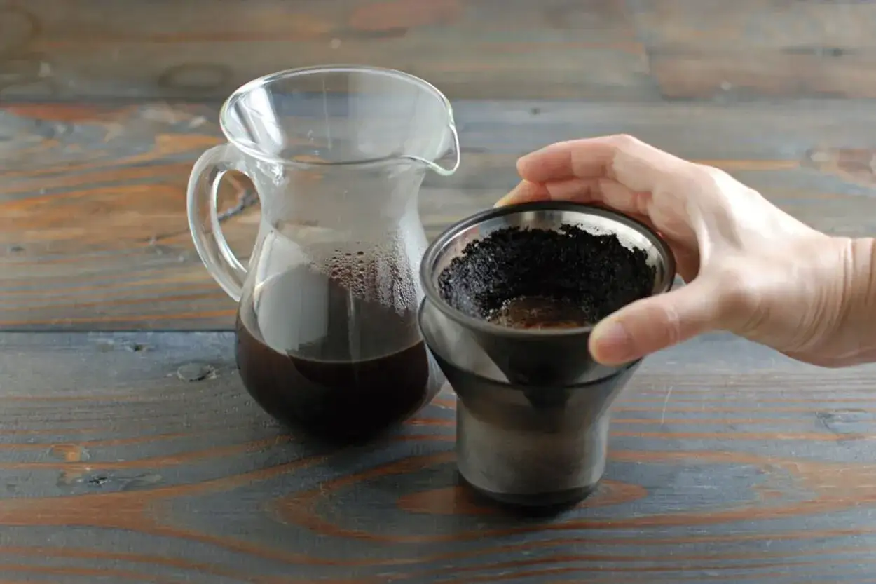 Eine Hand hält einen Filter mit Kaffeesatz, daneben eine Karaffe mit frisch gebrühtem Kaffee. So einfach ist es, Kaffee zu kochen.