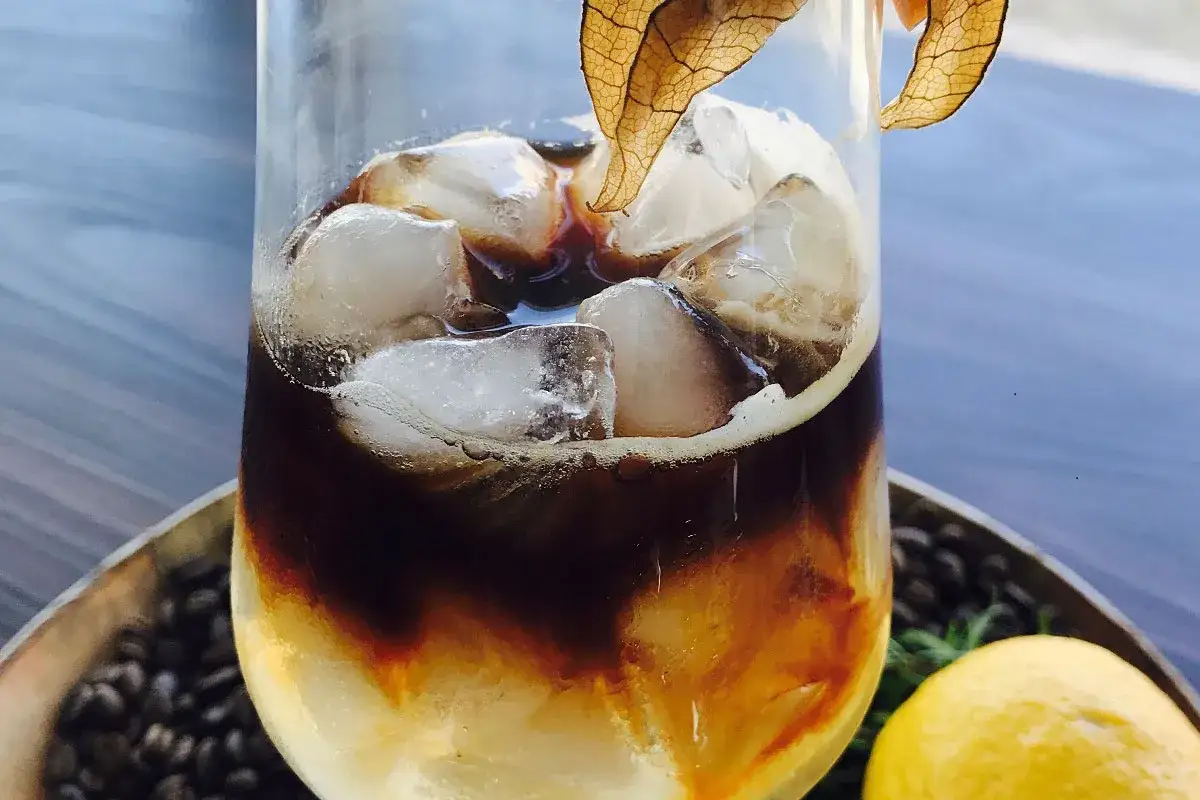 Erfrischender Espresso Tonic mit Eisw&uuml;rfeln, garniert mit einer Physalis. Ein Zitronen- und Kaffeebohnen-Dekor rundet das Bild ab.
