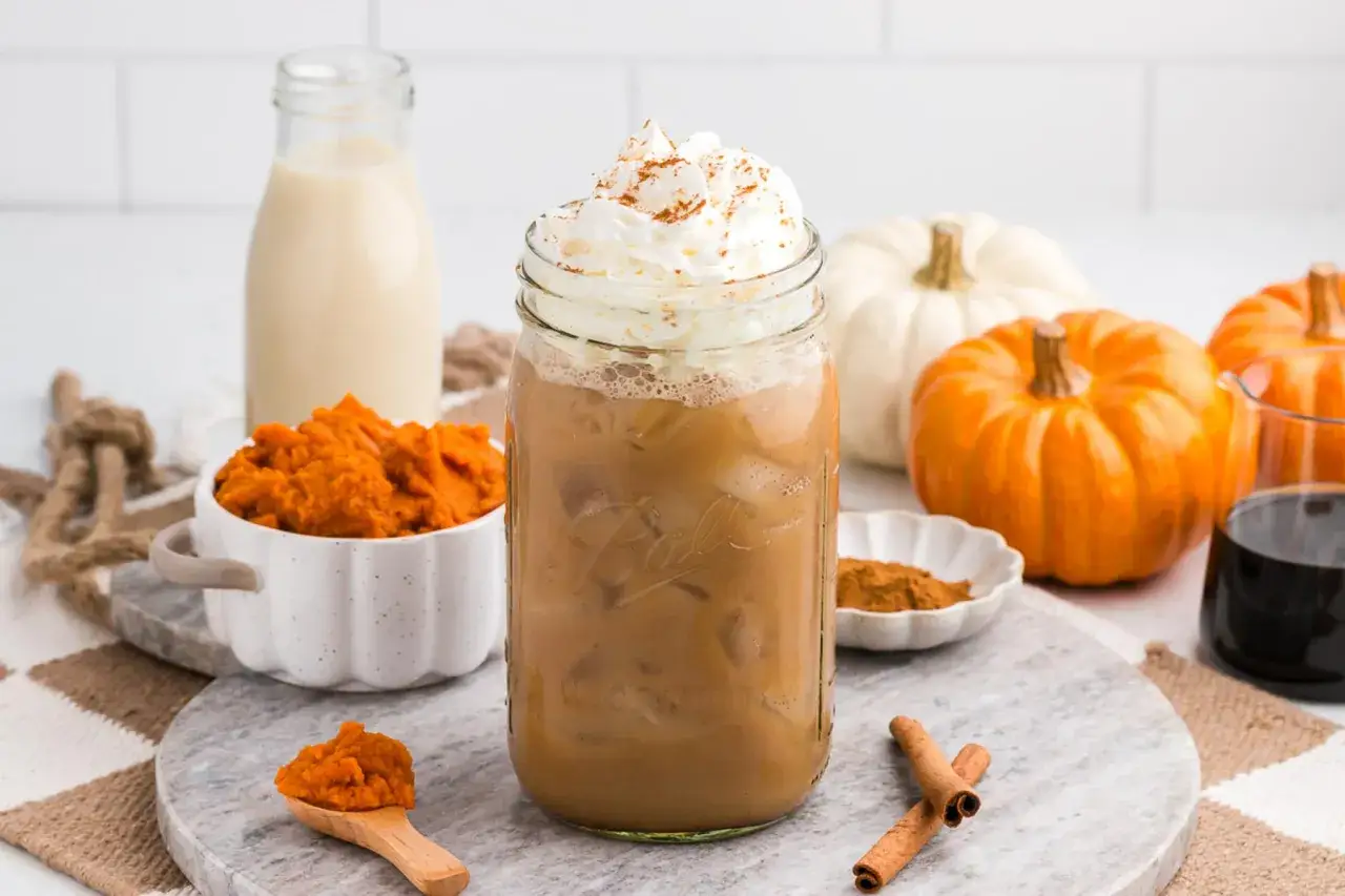 Eiskalter Pumpkin Spice Latte mit Sahne und Zimt, umgeben von Kürbissen und Zutaten für den perfekten Herbstgenuss.