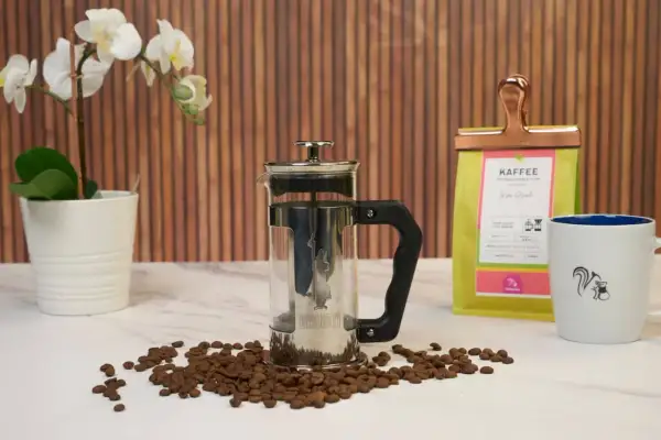 French press mit Kaffeesatz, bereit zum Entsorgen. Daneben Kaffeebohnen, eine Kaffeetüte und eine Tasse.
