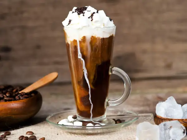 Ein erfrischender iced espresso mit Sahne, Marshmallows und Schokostreuseln, daneben Kaffeebohnen und Eiswürfel.
