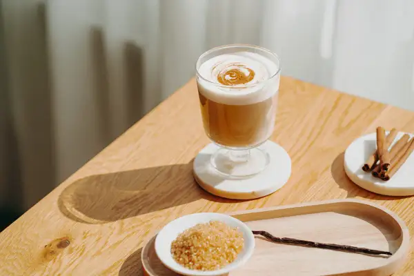 Ein cremiger Zimt Latte mit aufgeschäumter Milch und Zimtdekor. Daneben brauner Zucker, Zimtstangen und eine Vanilleschote.