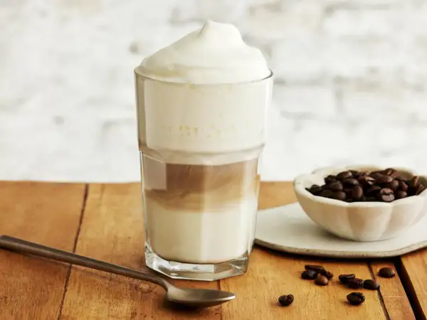 Ein hoher Latte Macchiato Eiskaffee mit Milchschaum, daneben eine Schale Kaffeebohnen und ein Löffel.