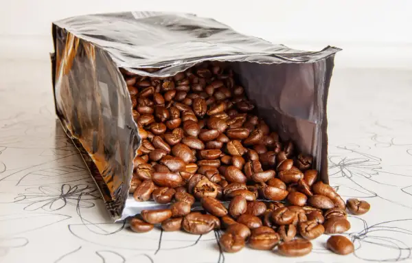 Ein offener Beutel mit gerösteten Kaffeebohnen, die auf eine gemusterte Oberfläche fallen. Die Frage, wie lange sind kaffeebohnen haltbar, beschäftigt jeden Kaffeeliebhaber.