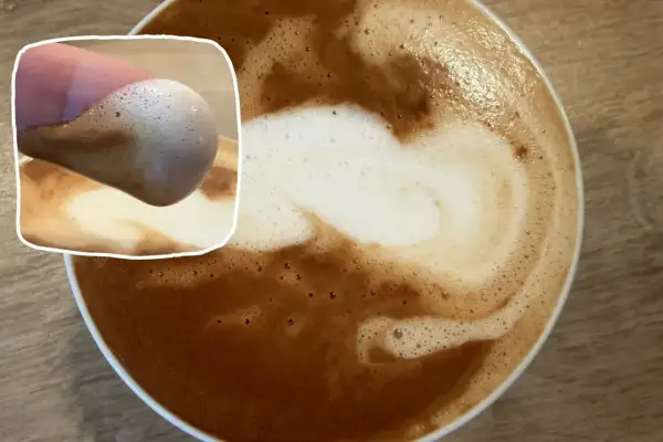 Ein Finger berührt den Schaum eines Kaffees mit Mandelmilch. Der cremige Schaum bildet ein Herzmuster.
