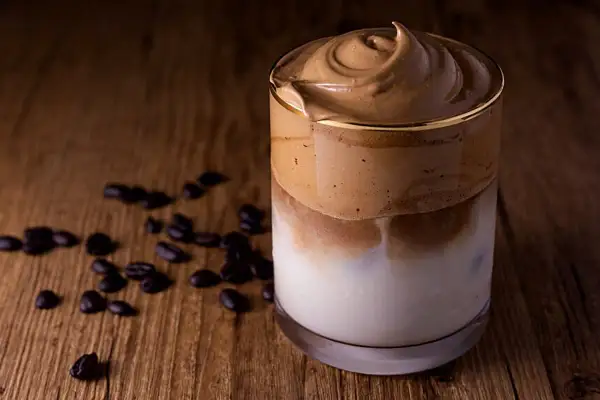 Ein cremiger Dalgona Kaffee ohne Zucker, serviert in einem Glas auf einem Holztisch, umgeben von Kaffeebohnen.
