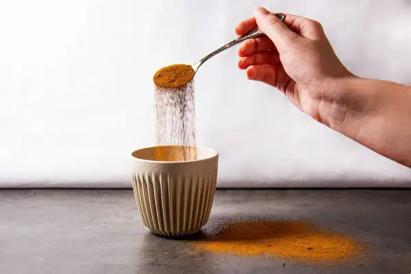 Eine Hand schüttet mit einem Löffel braunes Pulver in eine gerippte Tasse. Der Pulverregen erinnert an instant kaffee koffein.