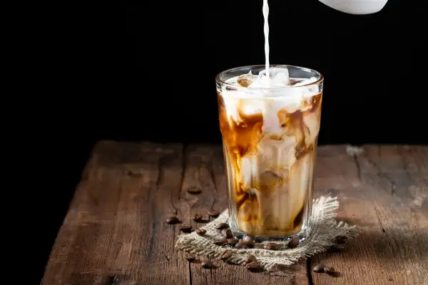 Milch wird in ein Glas mit Eis und Kaffee gegossen. Ein erfrischender Eiskaffee auf einem Holztisch.