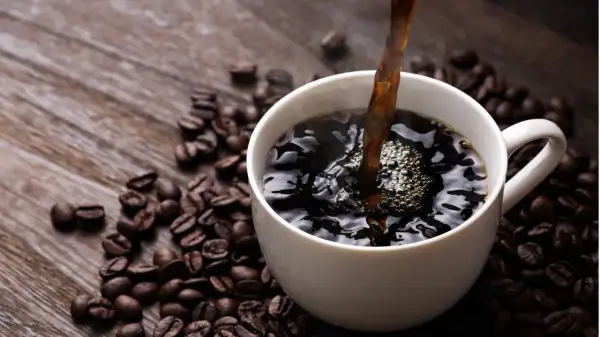 Heißer schwarzer Kaffee wird in eine weiße Tasse gegossen, umgeben von gerösteten Kaffeebohnen. Ein gesunder Start in den Tag.