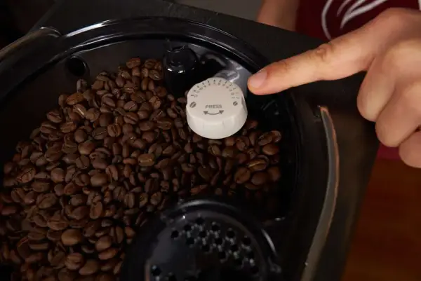 Finger stellt den Mahlgrad am Kaffeevollautomaten ein. Zahlreiche Kaffeebohnen sind im Behälter sichtbar.
