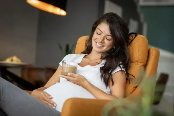 Glückliche Schwangere genießt einen Kaffee. Sie sitzt entspannt in einem Sessel und hält ein Glas mit einem cremigen Getränk.